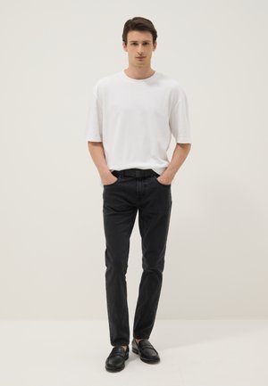 Junger Mann steht mit den Händen in den Taschen, trägt ein weißes T-Shirt, schwarze Jeans und schwarze Loafers vor einem einfarbigen beigen Hintergrund.