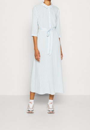 Frau trägt ein midi Hemdblusenkleid mit hellblauen und weißen vertikalen Streifen, dreiviertellangen Ärmeln und weißen Turnschuhen.