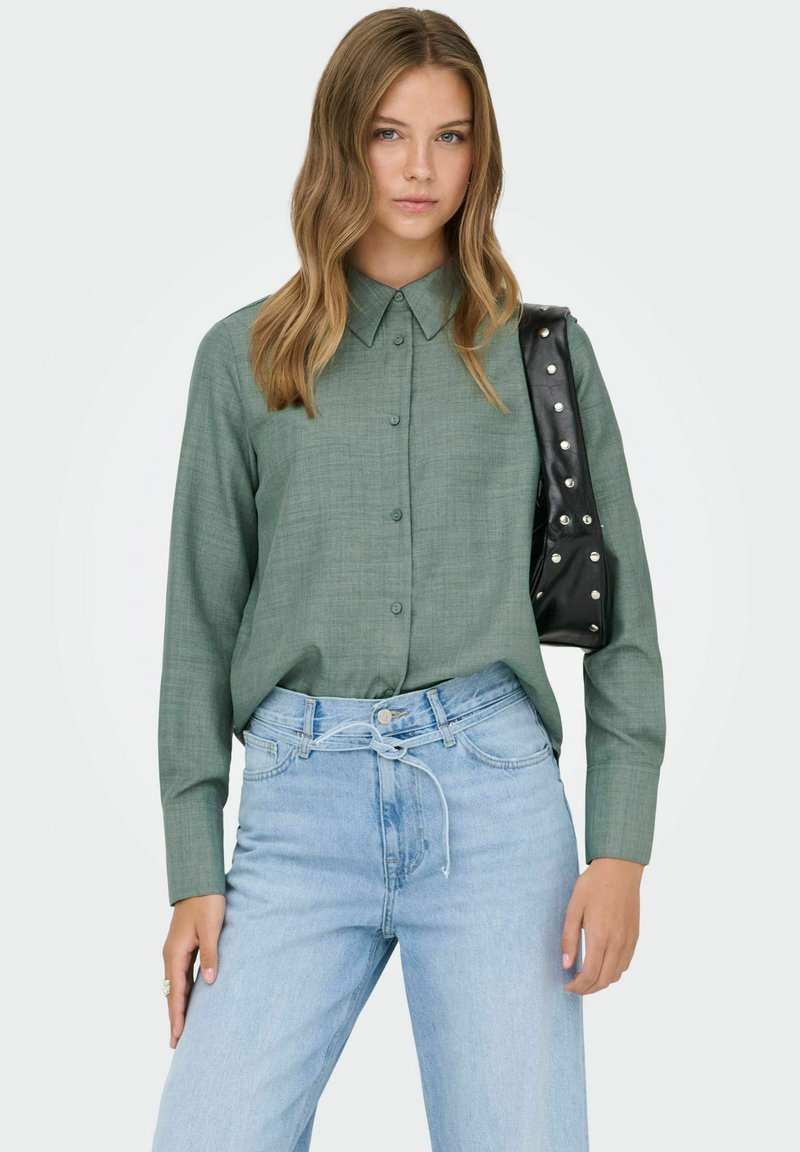 Camisa verde abotonada con cuello, combinada con vaqueros de talle alto en azul claro. Bolso negro con tachuelas plateadas colgado del hombro.