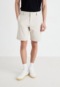 Beige shorts med rett snitt, liv med beltehemper, sidelommer og glatt tekstur, paret med hvite joggesko og sokker.