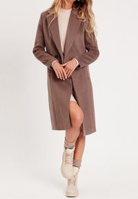Cappotto lungo marrone in lana con colletto a rever e chiusura a un bottone; indossato sopra un maglione chiaro e abbinato a stivali in crema con lacci.