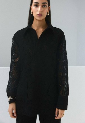 Mujer con blusa negra de encaje de manga larga, pendientes de aro dorados y brazaletes, de pie frente a una pared lisa gris y blanca.