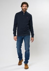 Pullover de forro polar azul marino con media cremallera y mangas largas, puños acanalados y un pequeño logo en el pecho. Combinado con unos vaqueros azules y botas marrones.