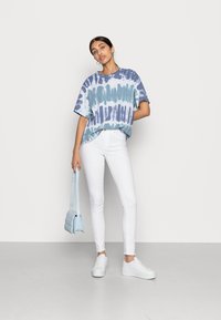 Sieviete valkā zilu un baltu t-kreklu ar krāsainu rakstu, baltas šaurās džinses, baltus sporta apavus, tur rokās gaiši zilu piešuvuma somu, stāvot pret vienkāršu fona.