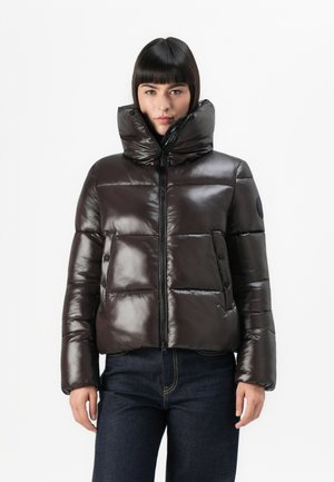 Vrouw in een glanzende donkerbruine pufferjack met een hoge kraag en een donkere jeans, staand tegen een effen witte achtergrond.