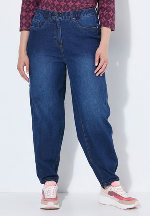 Person iført blå denimjeans med elastisk talje og pink-hvide sneakers står mod en hvid baggrund.