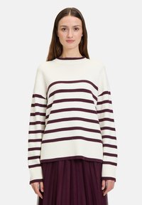 Pull en maille crème avec des rayures horizontales bordeaux, col rond, coupe ample et poignets côtelés. Associé à une jupe plissée noire.