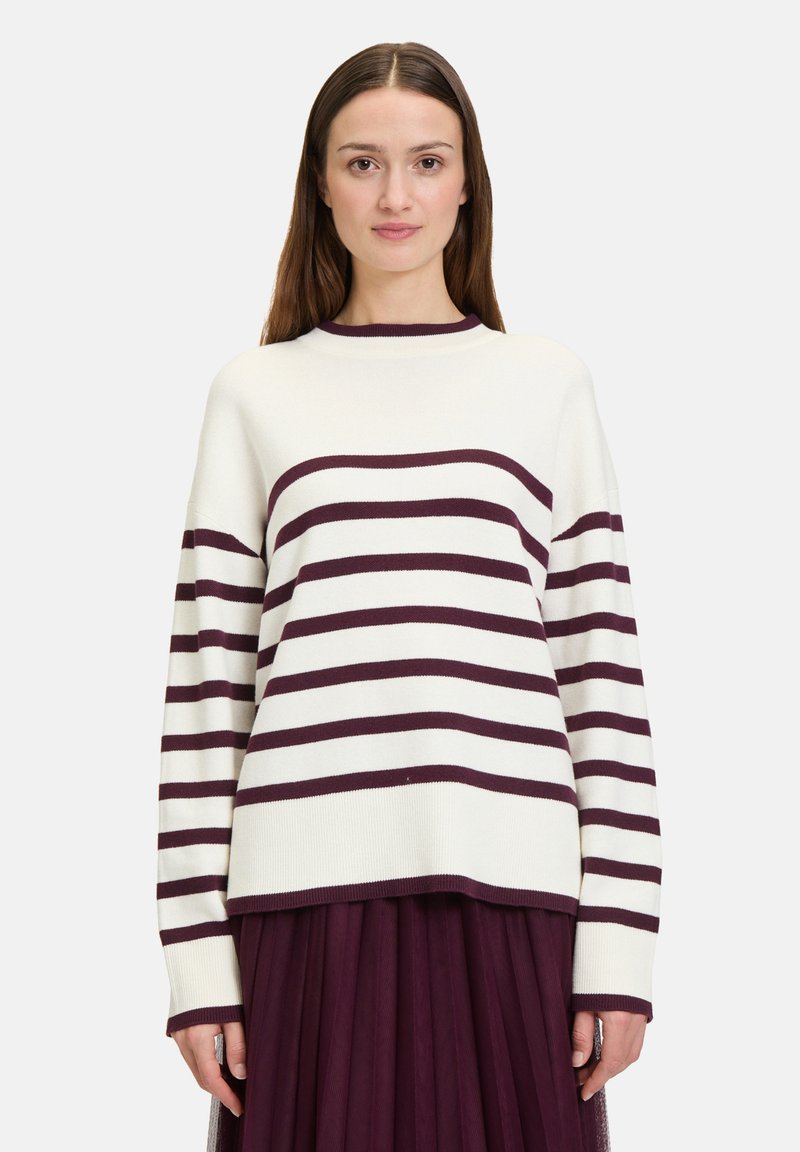 Pull en maille crème avec des rayures horizontales bordeaux, col rond, coupe ample et poignets côtelés. Associé à une jupe plissée noire.