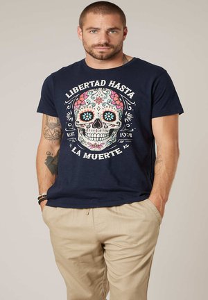 Homme avec barbe et tatouages portant un t-shirt bleu marine avec un crâne sucré coloré et un texte en espagnol, assorti à un pantalon beige, mains dans les poches.