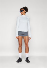 Hellblaues langärmeliges Zip-Up-Oberteil, graue Shorts, weiße Socken und schwarze Schuhe. Mit Daumenlöchern in den Ärmeln und reflektierendem Logodetail.