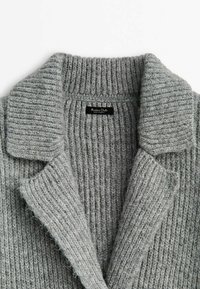 Cardigan en laine grise tricotée présentant une texture côtelée, un col châle et une étiquette cousue à l'intérieur. Matière douce et lisse avec un design décontracté.