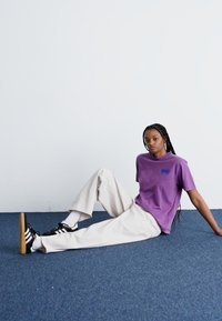 T-shirt en coton violet avec petit logo bleu, associé à un pantalon large beige clair et des baskets noires avec des rayures blanches.