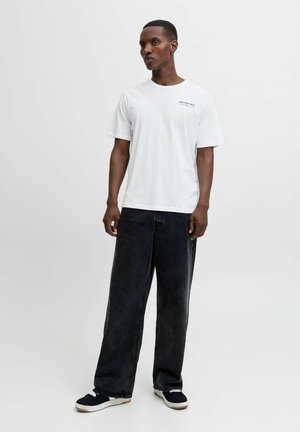 Homme debout, portant un t-shirt blanc avec un petit texte, un pantalon noir ample, des baskets noir et blanc, sur un fond clair uni.
