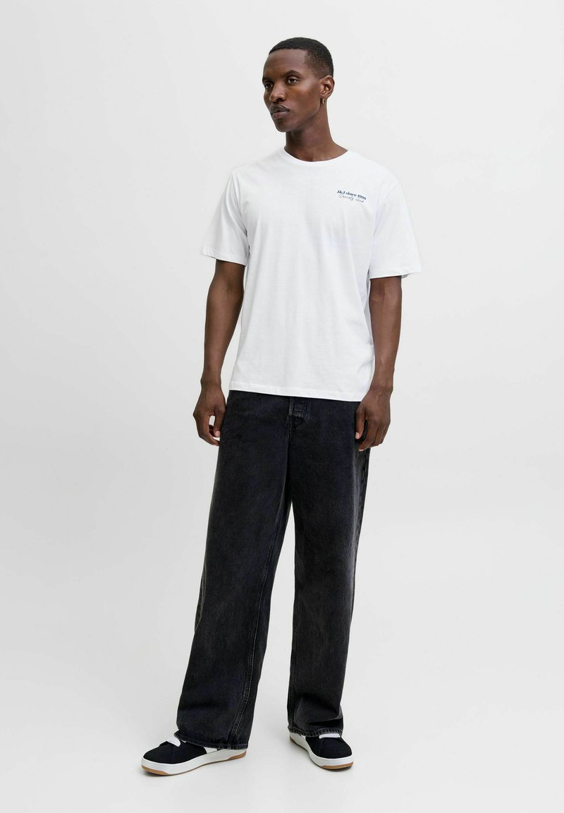 Hombre de pie, vistiendo una camiseta blanca con texto pequeño, jeans negros holgados, zapatillas en blanco y negro, fondo claro liso.