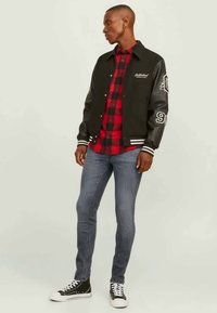 Juodas sportinis švarkas su odinėmis rankovėmis, juostelėmis ant rankovių, dėvimas su raudona ir juoda languota marškinėliais, mėlynomis džinsais ir juodais aukštakulniais sportbačiais.