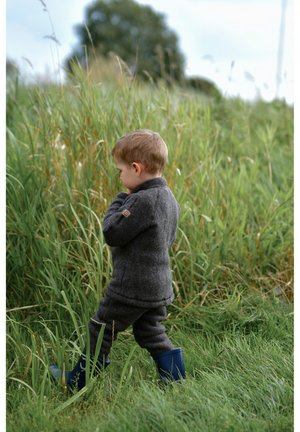 Bērns pelēkā vilnas džemperī un biksēs stāv garajā zaļajā zālē, valkājot zilas gumijas zābakus, skatoties prom no kameras.