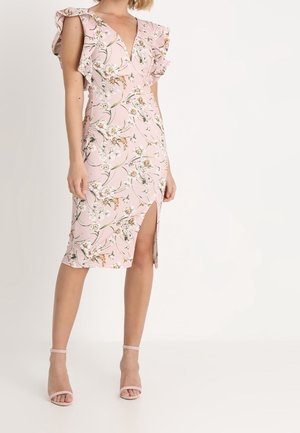 Femme portant une robe rose à fleurs avec un col en V, des manches à volants et une fente à l'avant de la cuisse, associée à des talons roses à bride à la cheville.