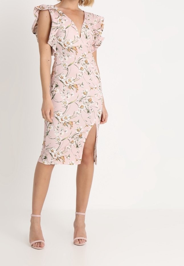 Femme portant une robe rose à fleurs avec un col en V, des manches à volants et une fente à l'avant de la cuisse, associée à des talons roses à bride à la cheville.