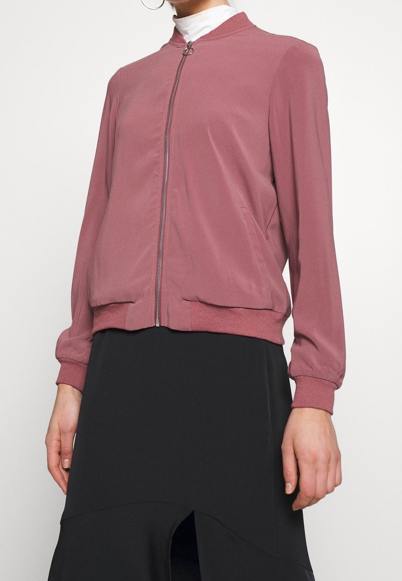 Veste bomber rose clair avec fermeture éclair à l'avant, col et poignets côtelés, présentant une texture de tissu lisse. Portée sur une jupe noire fendue.