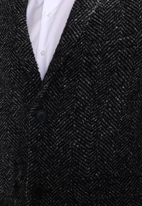 Svart tweedväst med fiskbensmönster och texturerad yta, försedd med två svarta knappar och en vit skjorta med krage under.