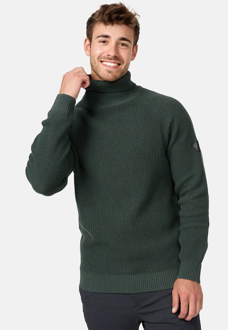 Zeleni turtleneck pulover s teksturom, s rebrastim rukavima i donjim rubom. Ima okruglu logotip oznaku na lijevoj rukavi i uski dizajn.