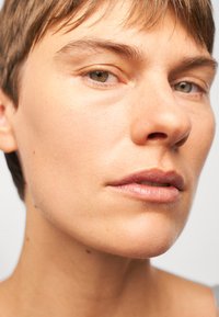 Close-up of a face with short hair, smooth light skin, defined cheekbones, and natural lips, against a soft, neutral background.