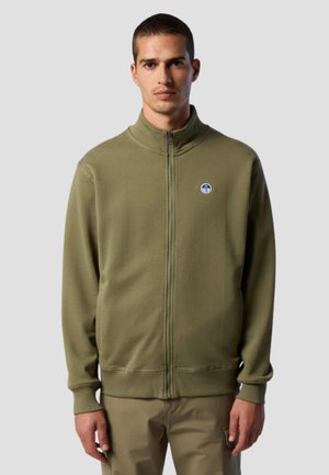 Man wearing olive green zip-up jacket with high collar and small round logo on left chest, standing against plain background.