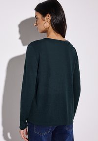 Tummanvihreä, pitkähihainen paita, jossa on pyöreä kaula-aukko ja sileä pinta, yhdistettynä tummiin farkkuihin. Takakuvassa näkyy siistejä linjoja ja minimaalinen muotoilu.