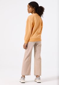 Sudadera naranja con puños acanalados, combinada con pantalones de pierna ancha color beige claro con dobladillos deshilachados. Lleva zapatillas grises y blancas.