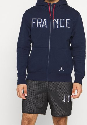Mann trägt einen marineblauen Kapuzenpullover mit Reißverschluss und dem Schriftzug "FRANKREICH" sowie schwarze Netzshorts mit dem Schriftzug "JORDAN" am linken Bein.