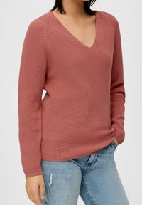 Rustfarbener Strickpullover mit V-Ausschnitt, gerippter Textur, langen Ärmeln und einem leicht lockeren Schnitt, kombiniert mit hellblauen, verwaschenen Jeans.