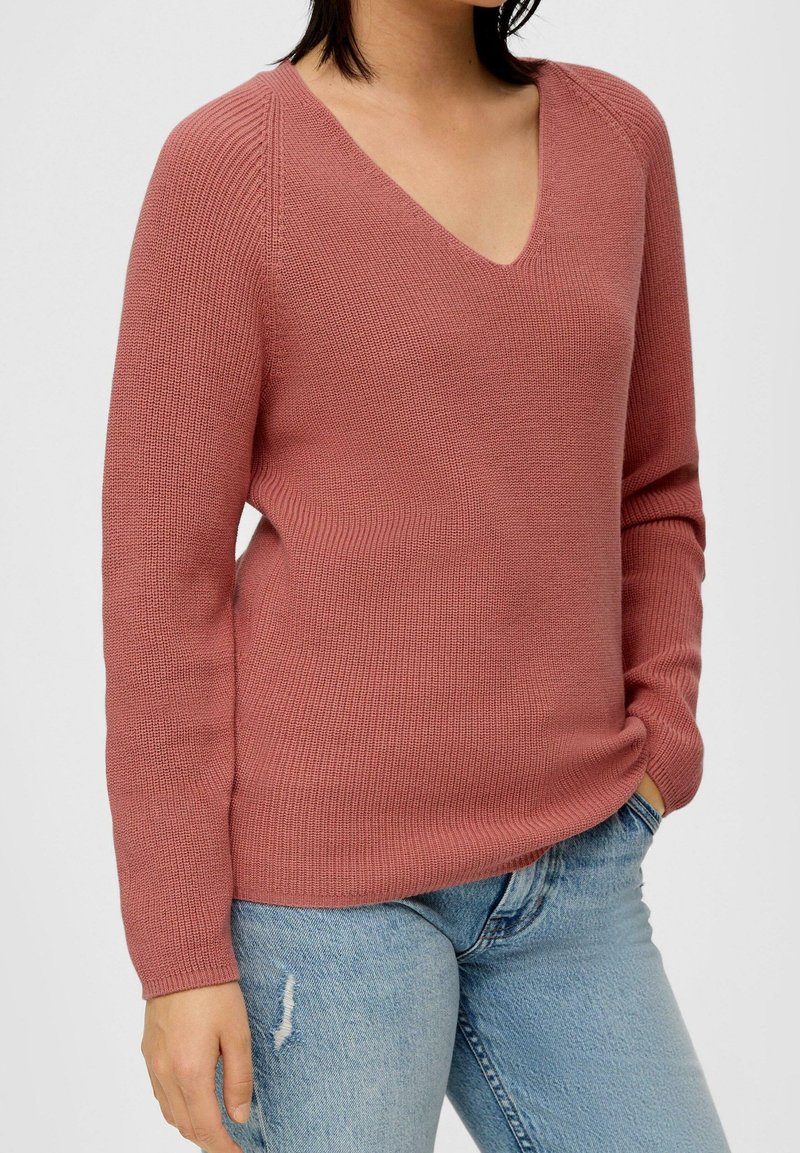 Rustfarbener Strickpullover mit V-Ausschnitt, gerippter Textur, langen Ärmeln und einem leicht lockeren Schnitt, kombiniert mit hellblauen, verwaschenen Jeans.