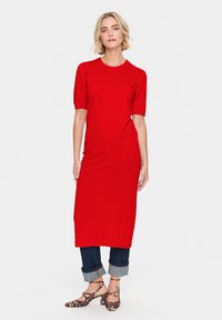 Abito in maglia rossa con maniche corte e silhouette aderente, abbinato a jeans scuri con risvolto e scarpe con cinturino alla caviglia a stampa leopardata.