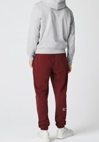 Sweat à capuche gris avec une poche kangourou, associé à un jogging bordeaux avec des poignets élastiques et un détail d'impression de texte blanc, porté avec des baskets blanches.