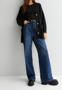 Černý pletený cardigan se stříbrnými knoflíky, v kombinaci s modrými širokými kalhotami s vysokým pasem a masivními černými platformovými botami.