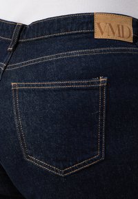Los jeans de mezclilla azul oscuro presentan un distintivo parche de cuero etiquetado como "VMD", costuras visibles y un diseño de un solo bolsillo trasero.