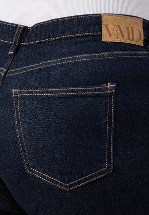 Ciemnoniebieskie jeansy z denimu z widocznym pomarańczowym przeszyciem i brązową skórzaną łatką z napisem „VMD” nad tylną prawą kieszenią.