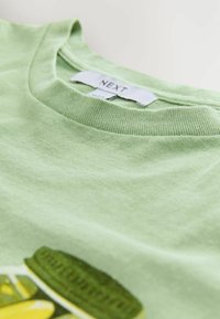 T-shirt en coton vert clair à col rond avec un tissage texturé, présentant un imprimé graphique aux couleurs vives près de l'ourlet. Étiquette de marque visible.