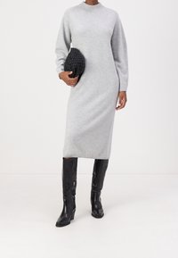 Robe en tricot grise avec col rond et manches longues, associée à des bottes à talons noires côtelées montantes jusqu'au genou et à une pochette noire texturée.