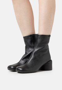Bottes en cuir noir mi-mollet avec fermeture éclair latérale et petit talon bloc, portées sur des jambes nues sur un fond clair uni.