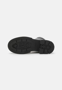Semelle de botte en caoutchouc noir avec un motif texturé, présentant un détail de logo et des zones de grip surélevées pour une traction optimale. Profil plat avec des bords arrondis.