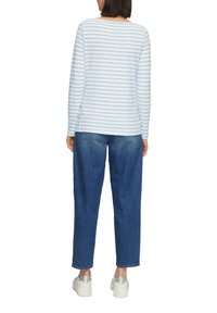 Osoba v modrobílé pruhované košili s dlouhým rukávem, modrých džínách a bílých platformových teniskách stojící zády ke kameře na bílém pozadí.