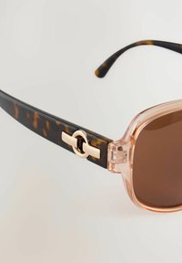 Lunettes de soleil avec une monture transparente de couleur rose clair et des branches en écaille de tortue, dotées d'un accent circulaire doré sur la charnière et de lentilles brunes.