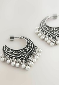 Sølvfarvede øreringe i hoop-design med indviklede blomster- og mønsterdetaljer, fremhævet med små metalperler langs nederste kant.