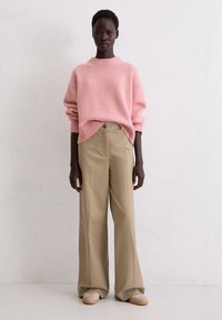 Helles rosa gestricktes Sweatshirt mit gerippten Details, kombiniert mit beigen, weit geschnittenen Hosen. Das Model steht vor einem neutralen Hintergrund. Braune Schuhe sind sichtbar.