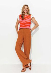 Débardeur au crochet à rayures rouges et roses, associé à un pantalon large marron. Les sandales présentent un design à lanières avec des clous.