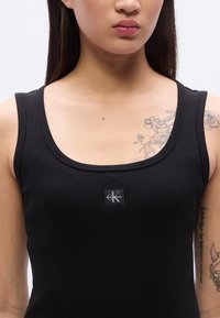 Μαύρο ριμπ τοπ με στρογγυλή λαιμόκοψη και μικρό λογότυπο Calvin Klein στο εμπρόσθιο μέρος. Ομαλή υφή υφάσματος και εφαρμοστή σιλουέτα.