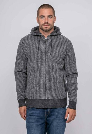 Homme aux cheveux courts portant un sweat à capuche zippé gris texturé et un jean bleu, debout sur un fond clair et uni.