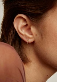 Pendientes de aro dorados con piedras multicolores, descansando en la oreja de un modelo con cabello castaño claro, mostrando una vista en primer plano.
