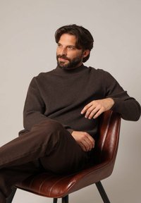 Pull côtelé marron avec un col montant, porté avec un pantalon marron foncé. Le mannequin est assis sur une chaise en cuir marron devant un fond neutre.